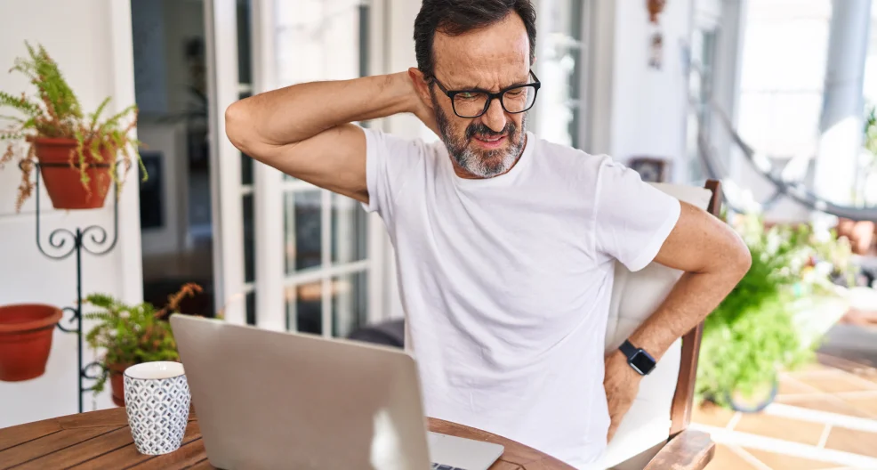 Middle age man using a laptop at home suffering of neck ache. He's touching his neck with his hand and seems to have discomfort.